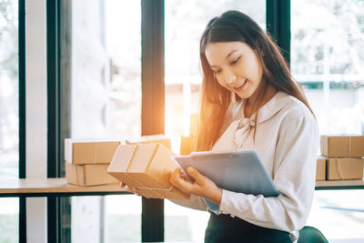 Smiling young woman holding glass while standing against window