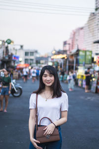 Portrait of young woman with purse standing on street in city