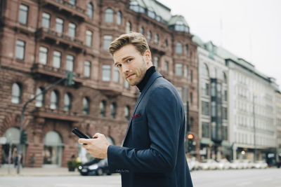 Portrait of businessman with smart phone standing against building in city