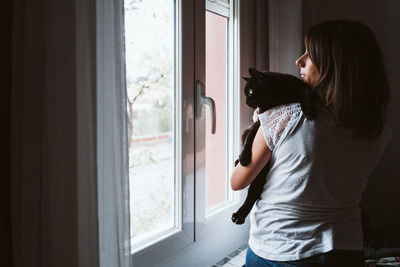 Side view of young woman looking through window at home