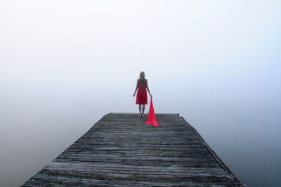 Rear view of woman holding scarf while standing on wooden pier by lake in foggy weather