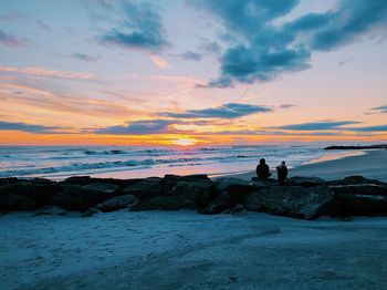 Rear view of people sitting on rocks at beach during sunset