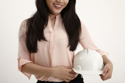 Midsection of engineer holding hardhat while standing against white background