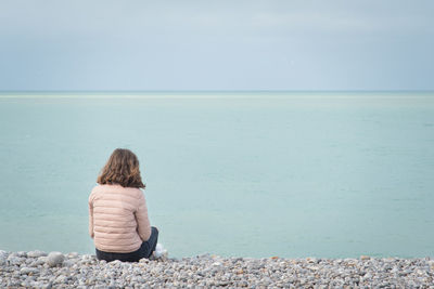 Rear view of teenage girl sitting at beach against cloudy sky