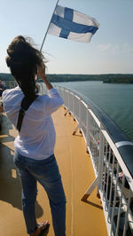 Rear view of woman holding flag while standing on boat deck in sea against sky