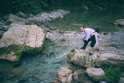 Man with camera looking away while standing on rocks by river