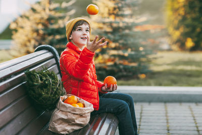 A boy in a bright orange jacket and a fashionable yellow hat sits on a bench, laughs, throws oranges 