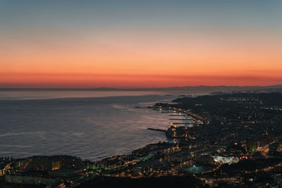 High angle view of illuminated city by sea against sky during sunset