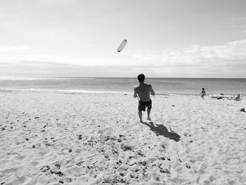 Rear view of shirtless man playing with kite at beach during sunny day