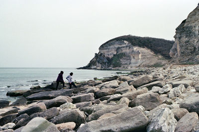 People looking at rocks by sea against sky