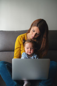 Busy mother hugging cute toddler daughter while sitting on sofa and doing remote project on laptop at home