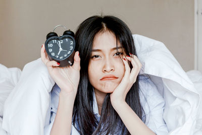 Portrait of sad woman showing alarm clock while lying on bed