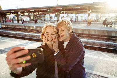 Cheerful young couple taking selfie through mobile phone on railroad station platform