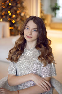 Teenage girl is sitting on a chair against the  of a christmas tree at home in a sequins dress