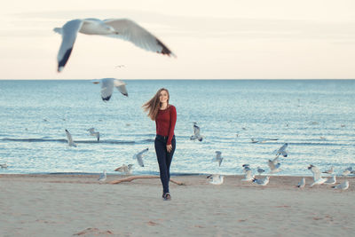 Full length portrait of beautiful young woman walking by birds at beach