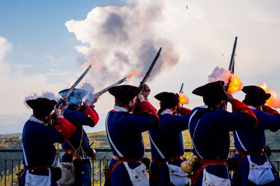 Rear view of army soldiers firing guns towards cloudy sky