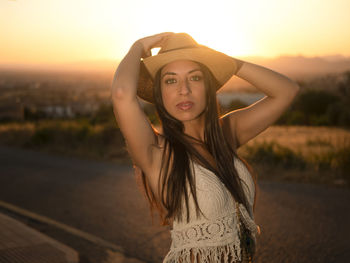 Portrait of young woman wearing hat standing on road at sunset