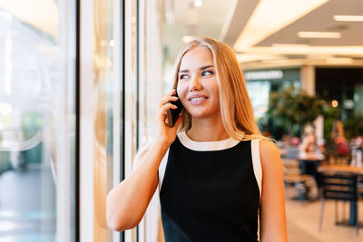 Cheerful female manager talking on cellphone and looking away with smile while standing near window of modern mall in daytime