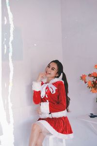 Smiling young woman wearing santa costume sitting against wall