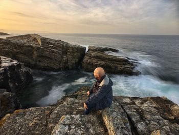 Senior man sitting on rock by sea against sky during sunset