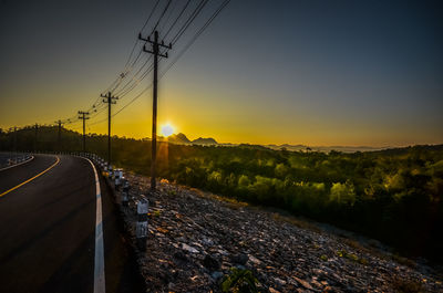 Road by electricity pylon against sky during sunset