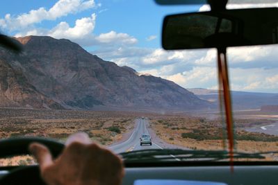 Cropped image of man driving car on country road leading towards rocky mountains