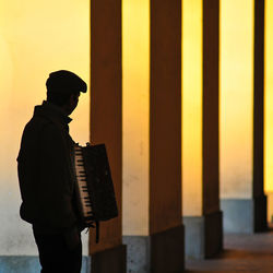Silhouette of a solitary accordion player street musician against a backdrop of yellow columns.