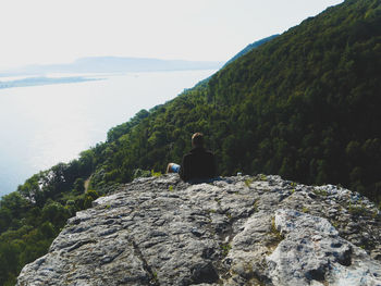 Rear view of man sitting on cliff by sea against clear sky