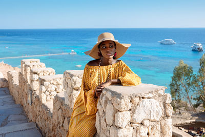 Happy african american woman in yellow dress and sun hat enjoys view of coast of red sea on natural 