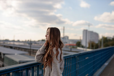 Young woman with eyes closed standing by railing against sky in city