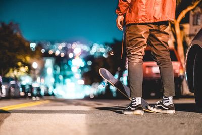 Low section of man with skateboard standing on road at night