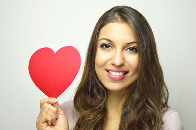 Portrait of beautiful young woman holding heart shape paper against gray background