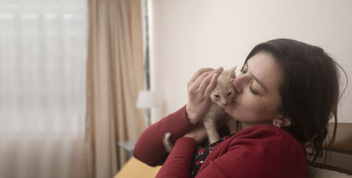 Portrait of hispanic young woman with red jacket hugging and kissing her pet kitten inside her room