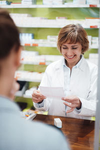 Smiling pharmacist reading prescription while standing with customer in store