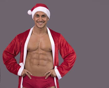 Portrait of smiling muscular man wearing santa hat while standing against gray background