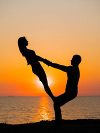 Silhouette couple exercising at beach against sky during sunset