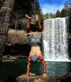 Full length of shirtless man performing handstand against waterfall