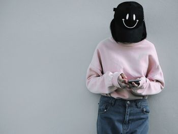 Woman wearing smiley cap using mobile phone against wall