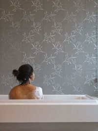 Rear view of shirtless woman bathing in tub against pattern wall