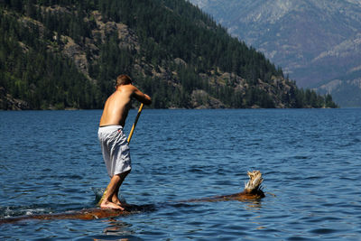 Full length of shirtless man paddleboarding in sea against tree mountains