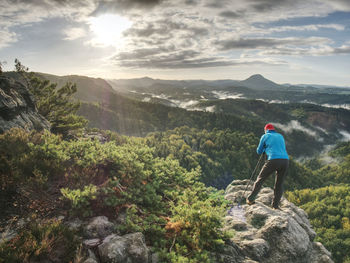 Photographer in blue sweatshirt working with mirror camera and tripod on peak of rock.