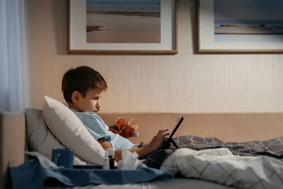 Little boy using tablet while laying in bed in living room. medicines and thermometer in foreground