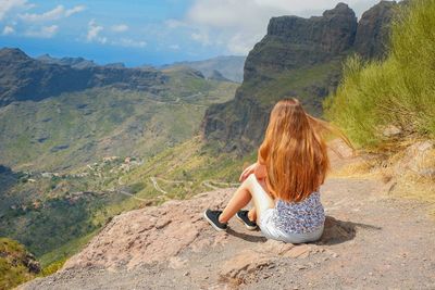 Rear view of young woman sitting on rock against sky