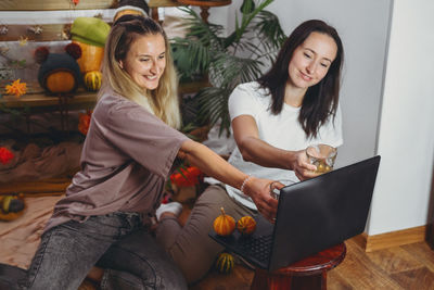Smiling women holding drinks while video calling on laptop at home