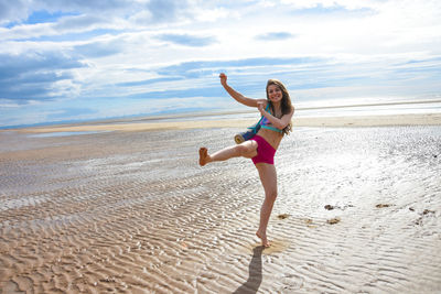 Full length portrait of young woman jumping at beach against sky