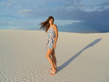 Full length portrait of beautiful woman standing on sand at beach against cloudy sky