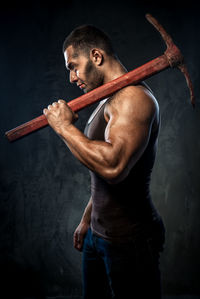 Side view of muscular man holding pick axe against wall