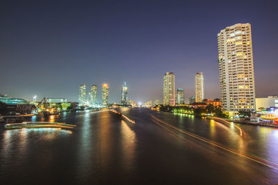 Light trails in river by illuminated city against clear sky at dusk