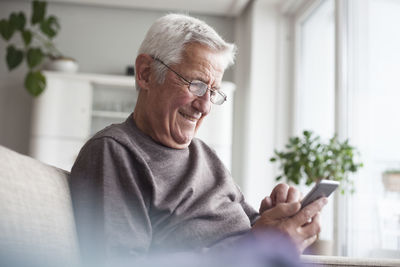 Portrait of smiling senior man sitting on couch at home using smartphone