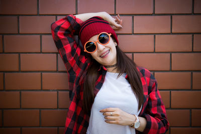 Portrait of smiling woman in sunglasses and knit hat standing against brick wall